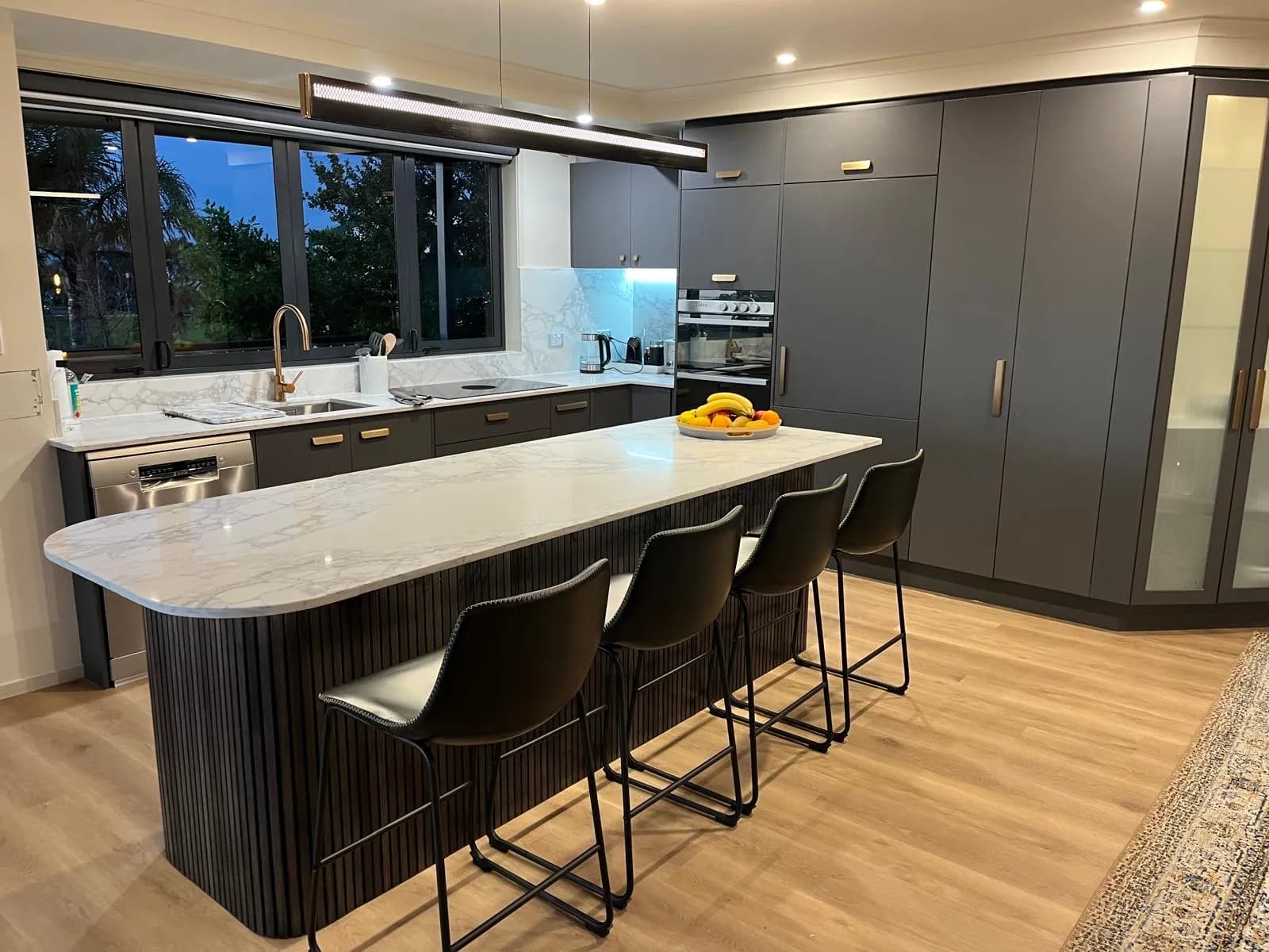 Curved fluted island with bar stools in front of charcoal tall cabinets at dusk