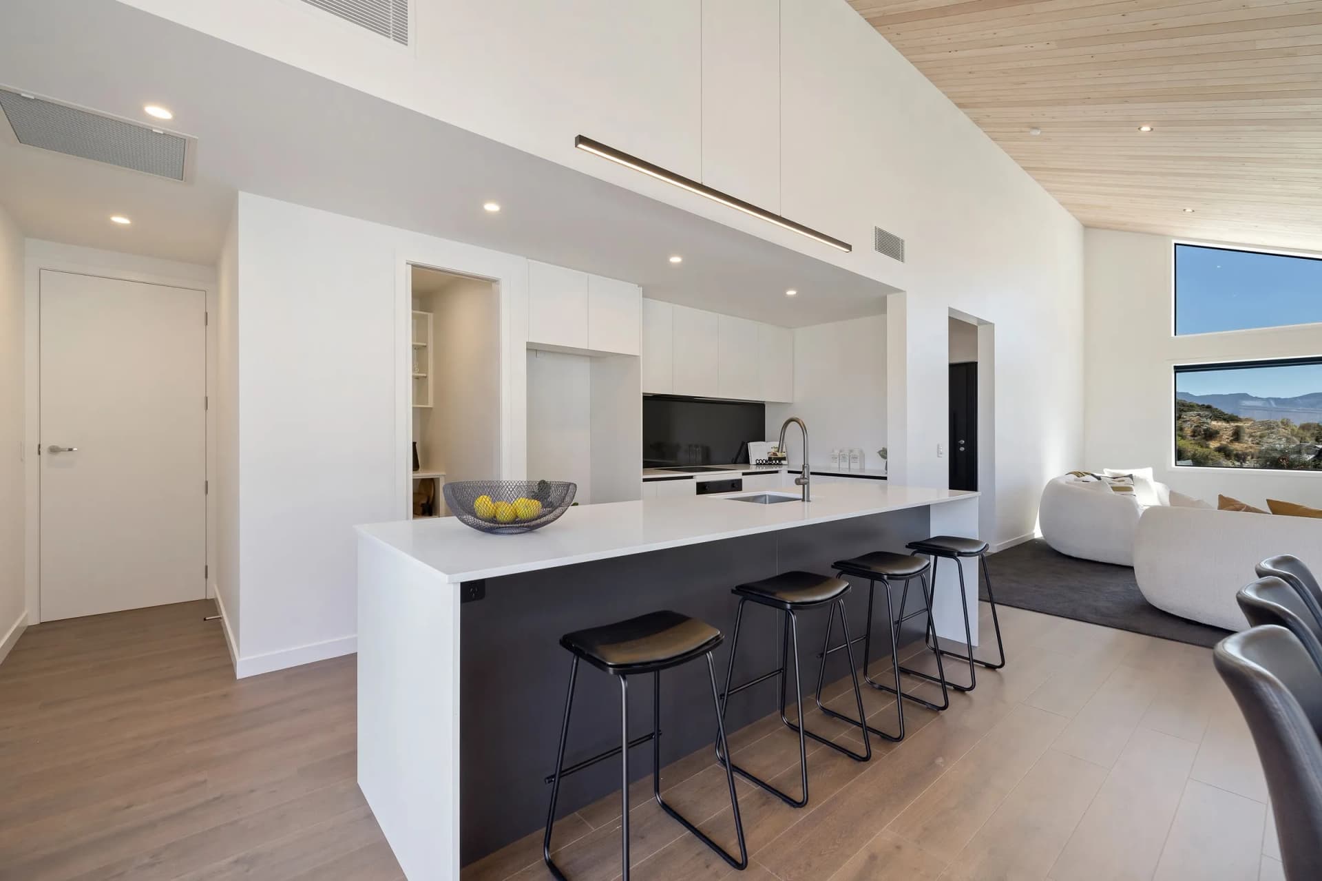 Dark island with a white waterfall benchtop and bar stools under a timber-lined ceiling