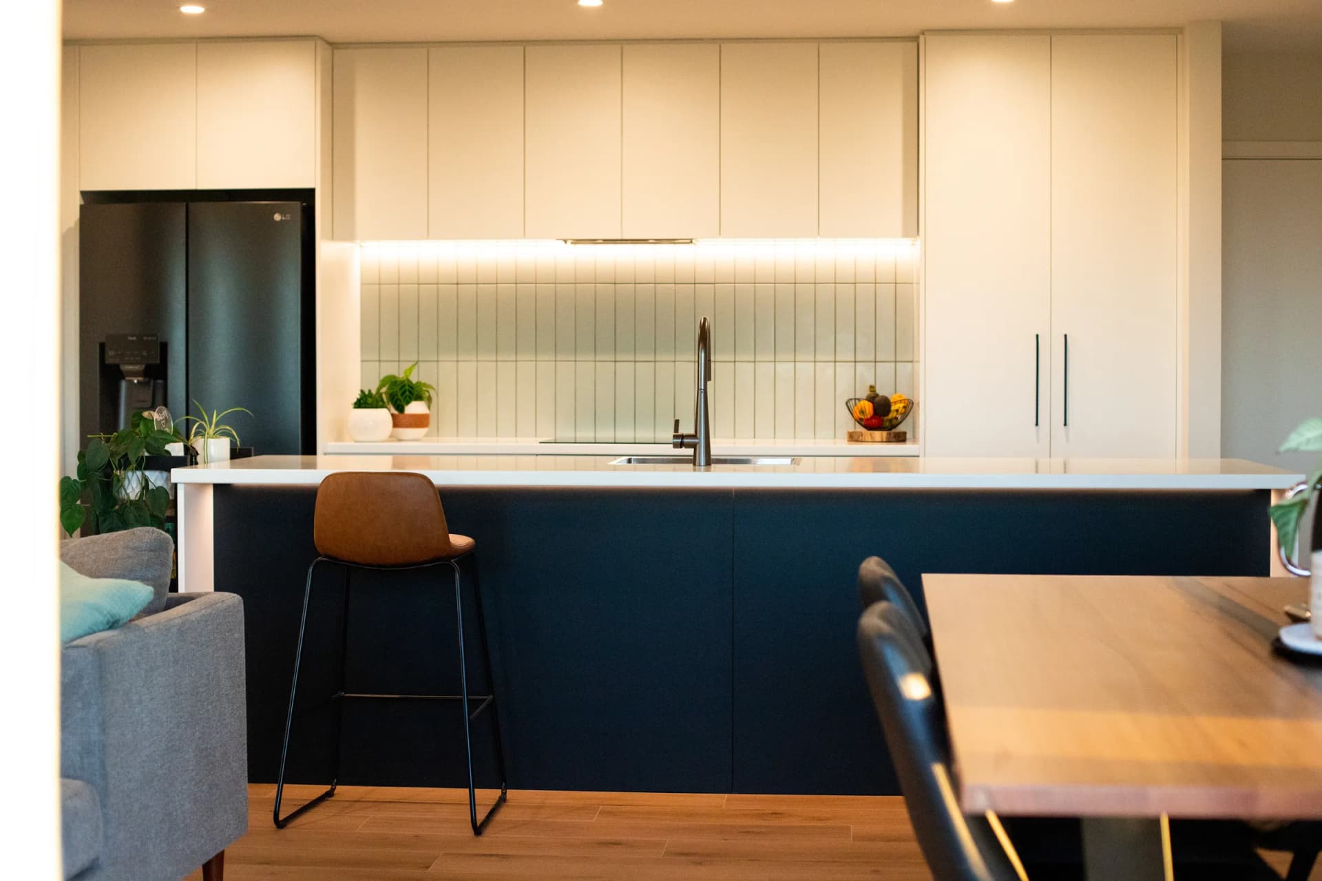Navy island with a stool in front of a white kitchen with a lit tiled splashback