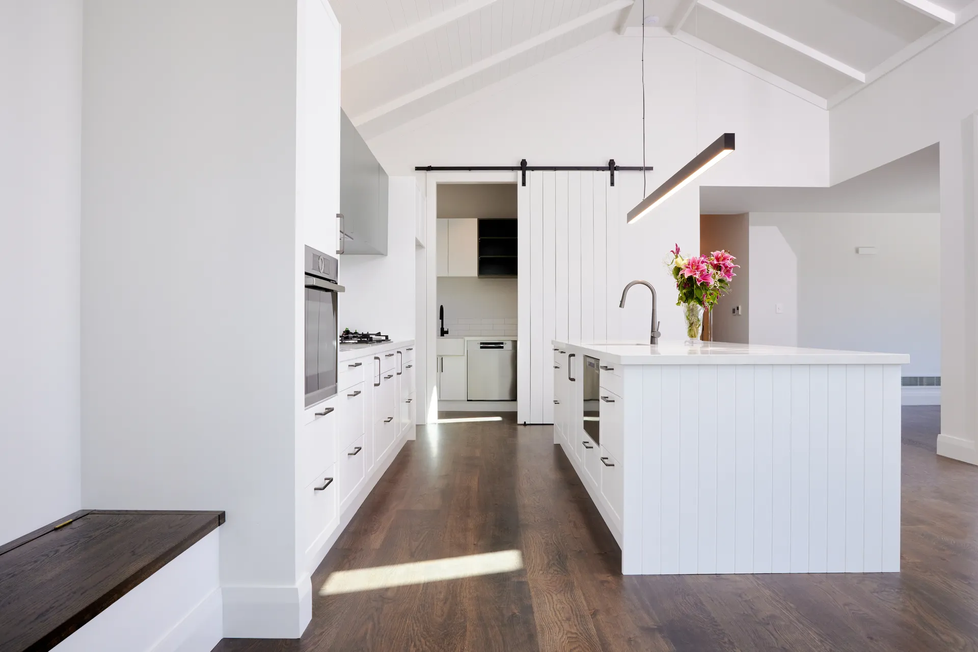 White panelled island and run of cabinets under a raked ceiling with a barn door beyond