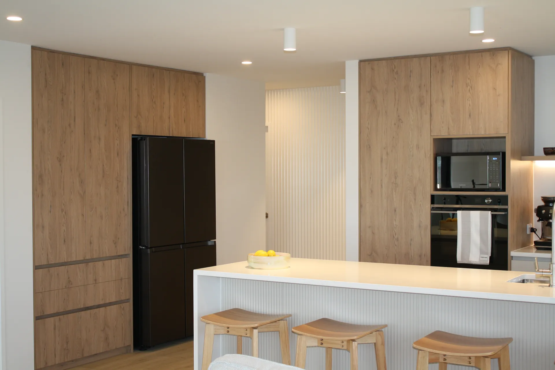 Black fridge set into timber-look tall cabinets beside a fluted white island