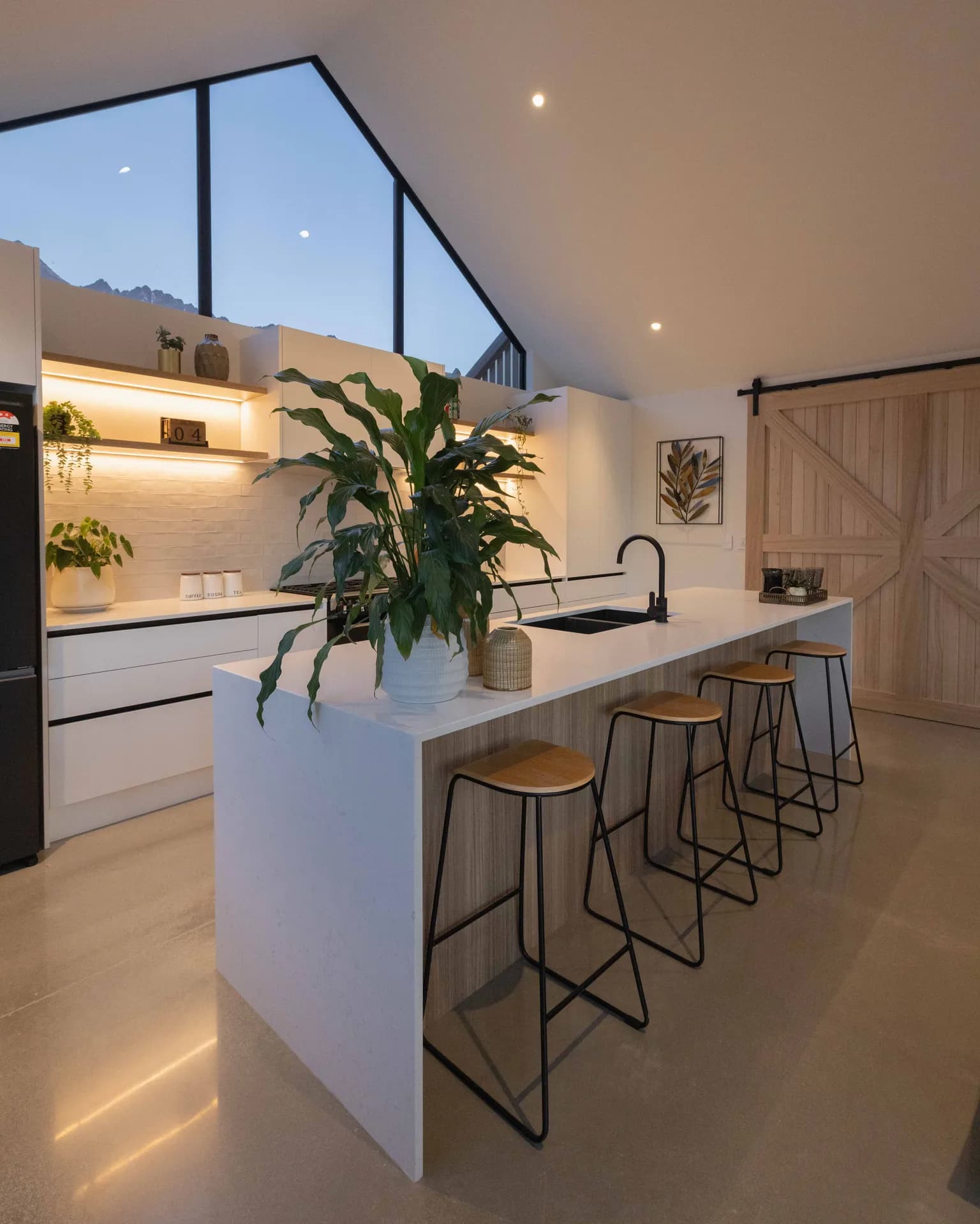 White waterfall island end with stools in front of lit open shelving under a gable window