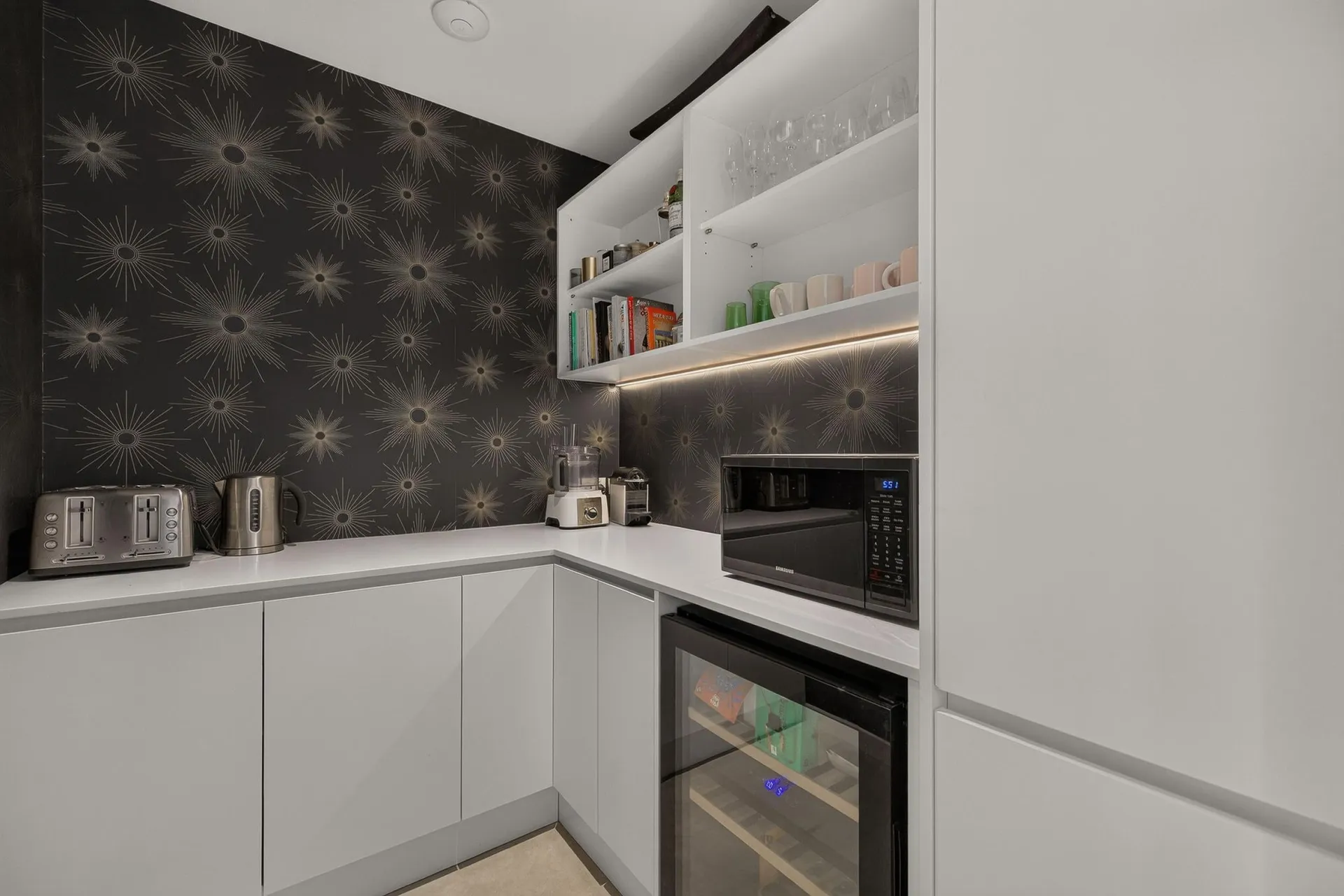 White scullery with lit open shelving, a wine fridge and patterned feature wallpaper
