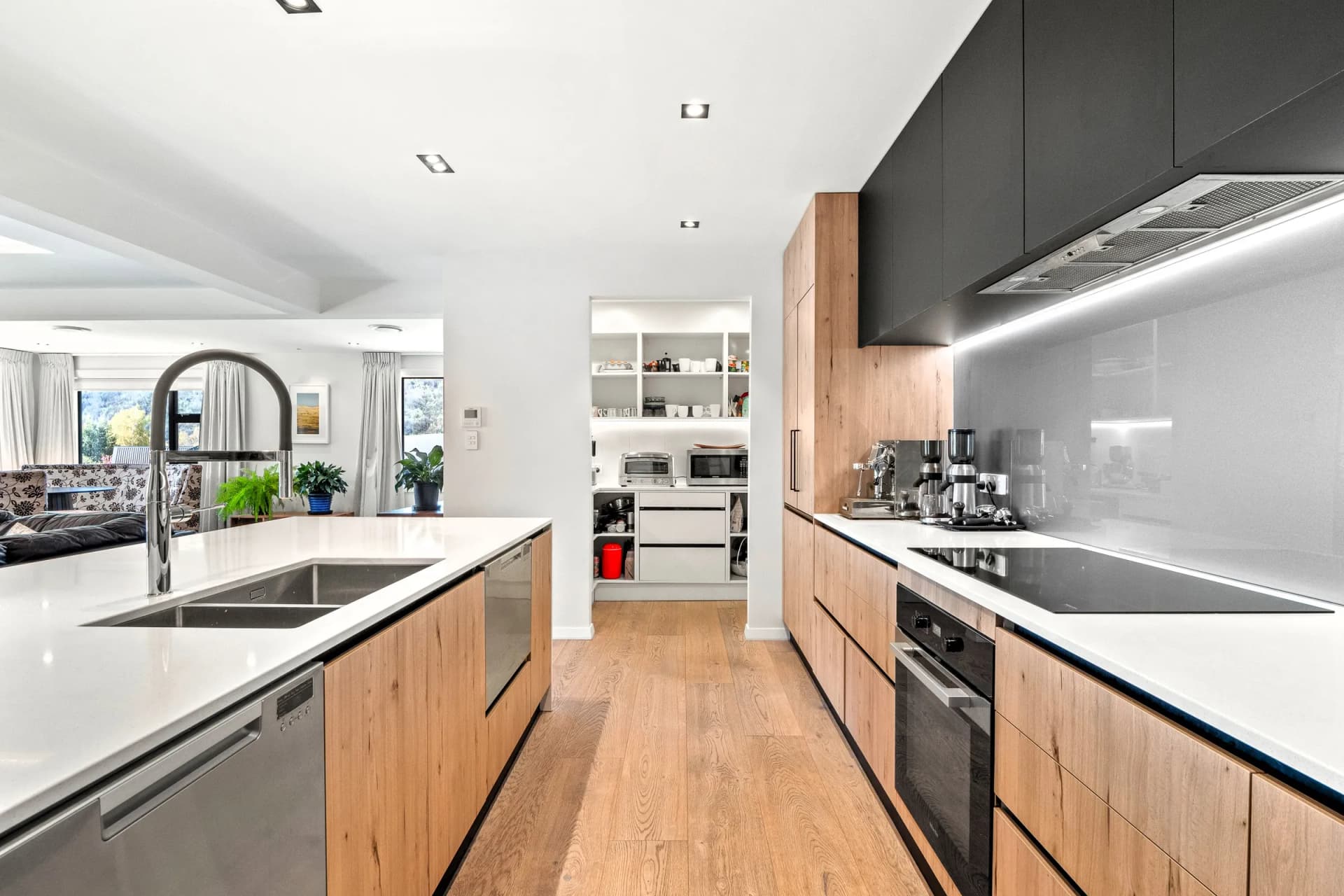 Galley between a white island and timber-look base cabinets under black overheads with a pantry beyond