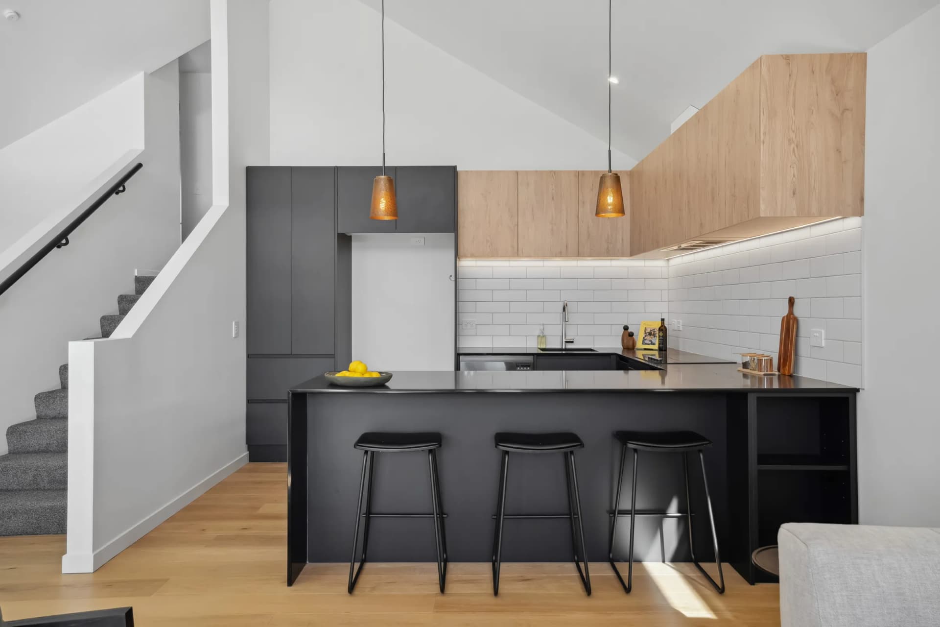 Black breakfast bar with stools below timber-look overheads and a white tiled splashback