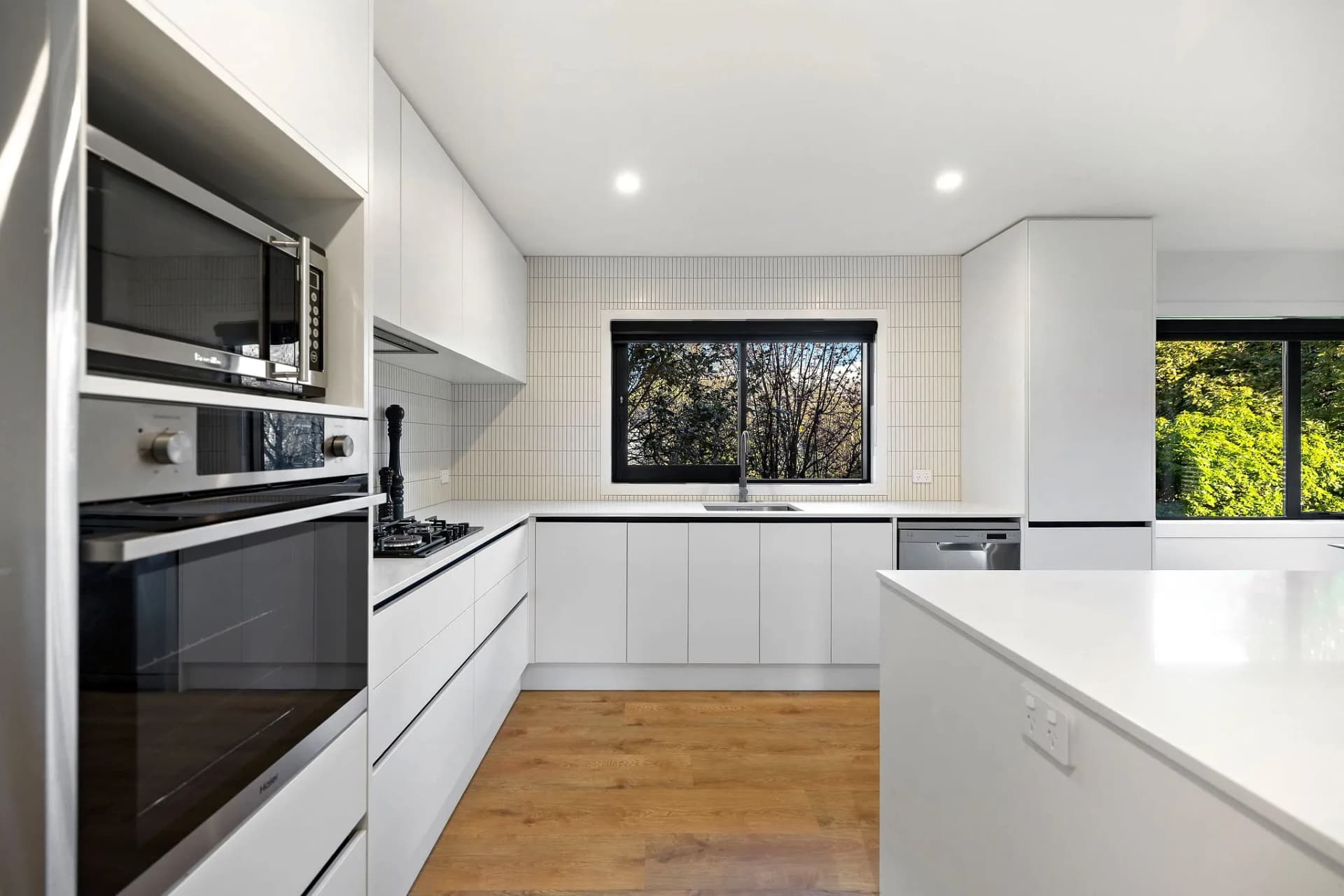 White handleless kitchen with wall ovens and a square tile splashback under a window