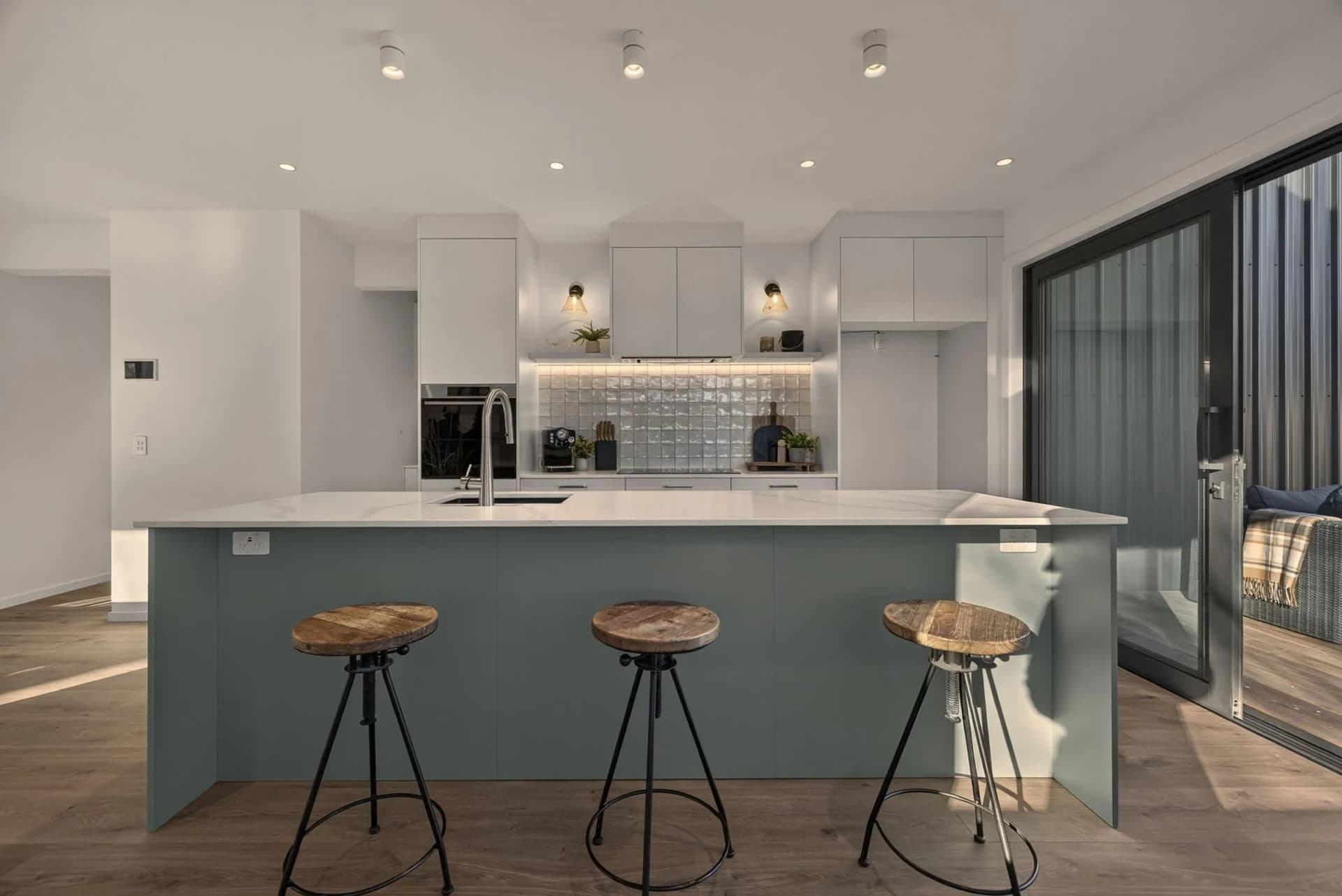 Grey-green island with three timber-seat stools in front of a white kitchen with a textured tile splashback