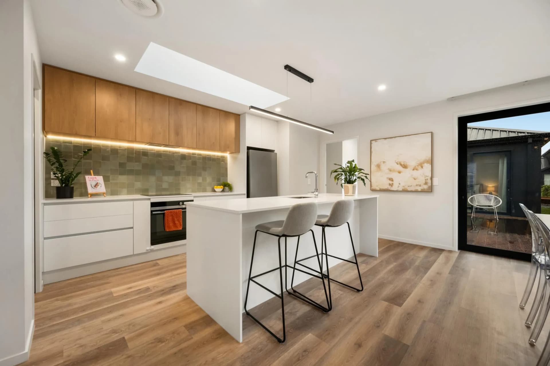 White island with bar stools below timber-look overheads, a green tiled splashback and a skylight
