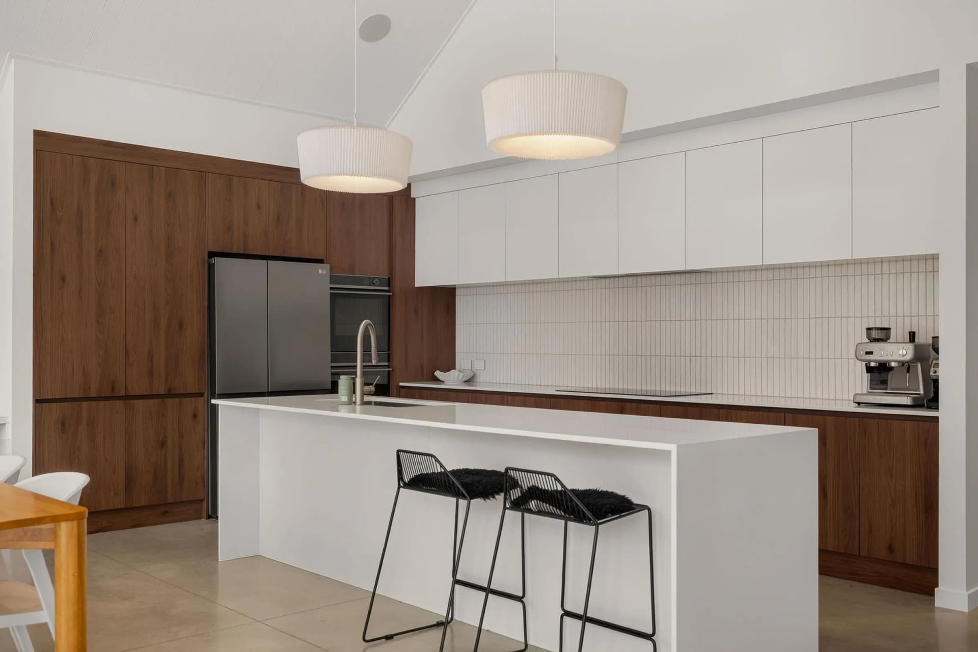White waterfall island with bar stools in front of tall timber-look cabinets and a white tiled splashback