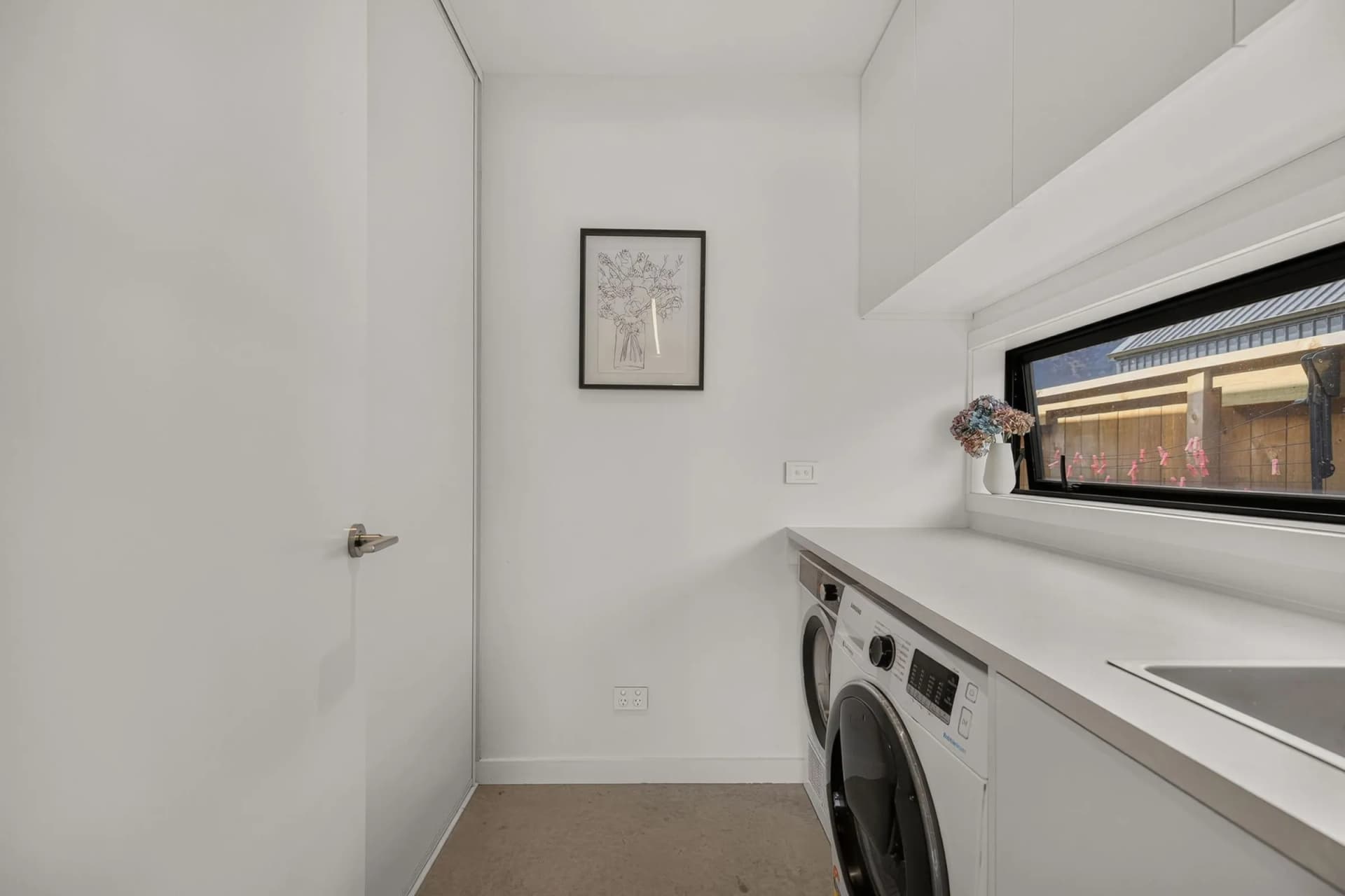 White laundry bench with a front-loader below overhead cupboards and a window