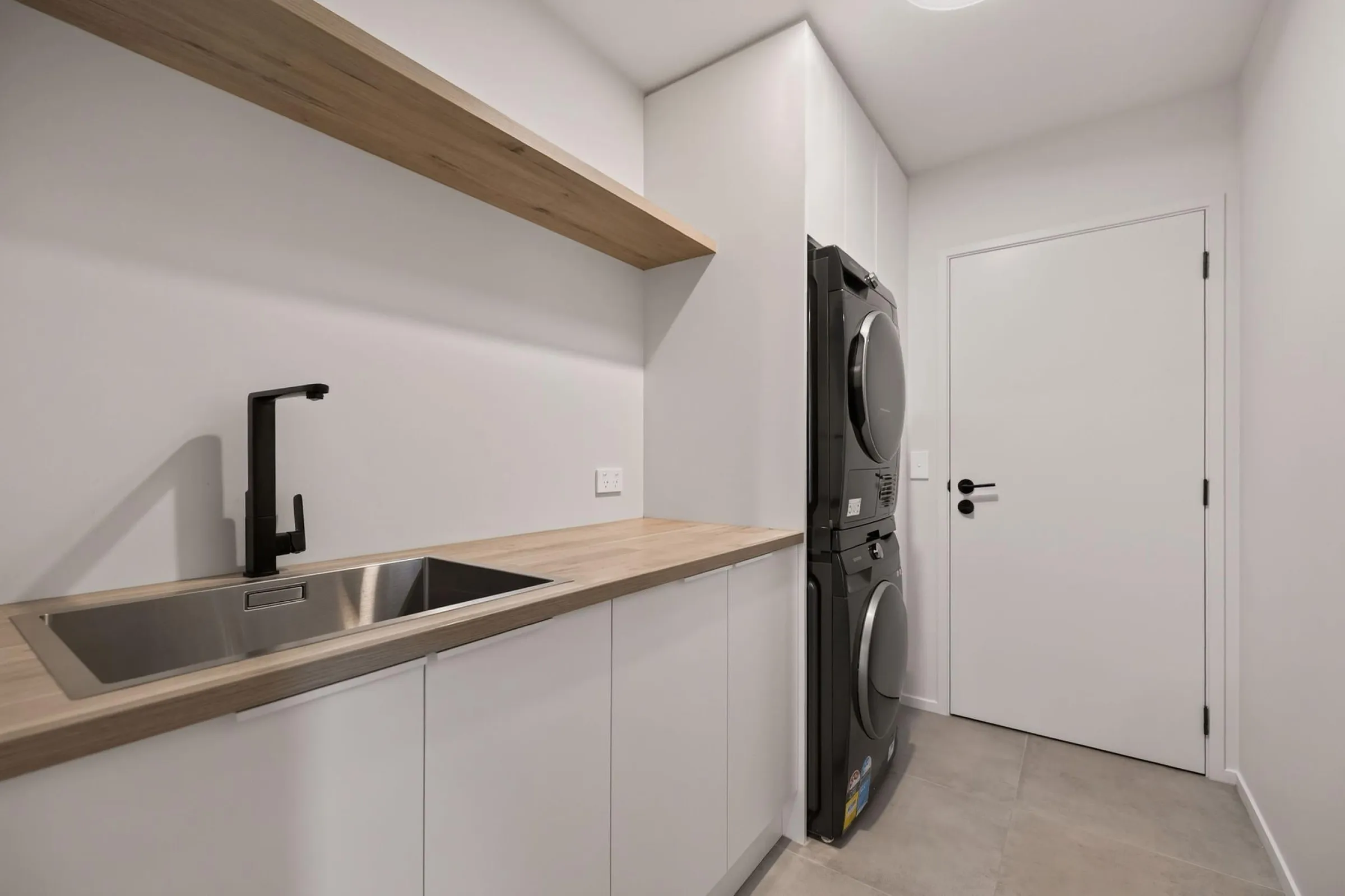 White laundry cupboards with a timber-look benchtop and shelf beside a stacked washer and dryer
